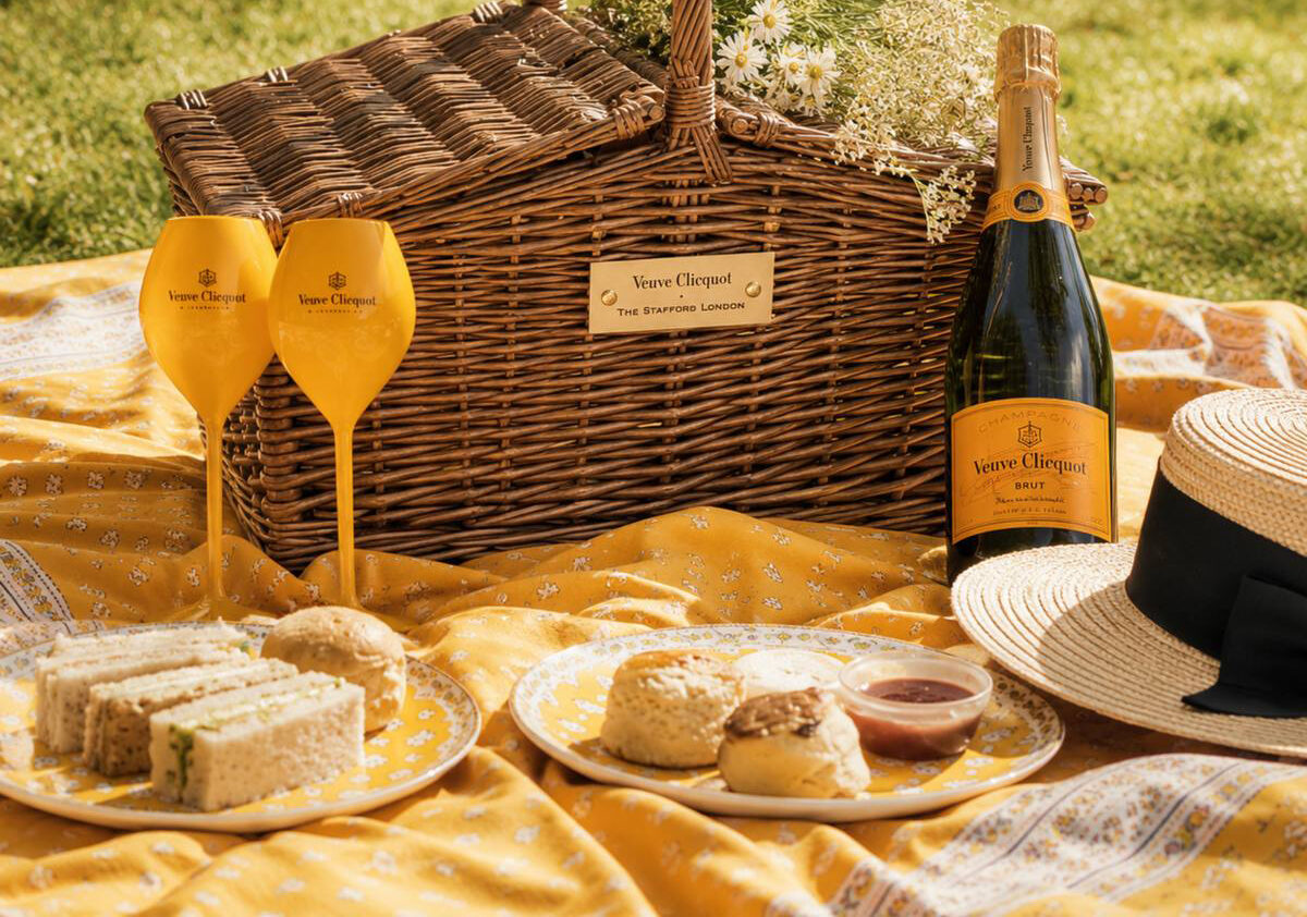 A wicker picnic basket with white flowers, two Veuve Clicquot champagne glasses, a bottle of Veuve Clicquot, and an array of treats are set on a yellow patterned blanket outdoors—perfect for a Chelsea Flower Show-inspired afternoon.