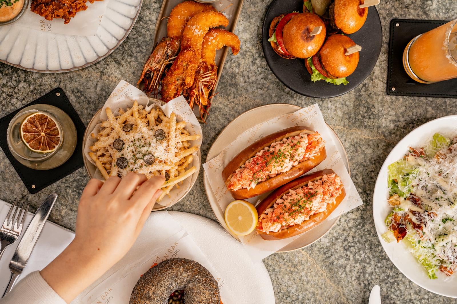 A table filled with various dishes, including lobster rolls, truffle fries, shrimp, mini burgers, a salad, a bagel, and drinks at the Enemigo Tequila Courtyard. A hand reaches for fries in the bottom left of the image.