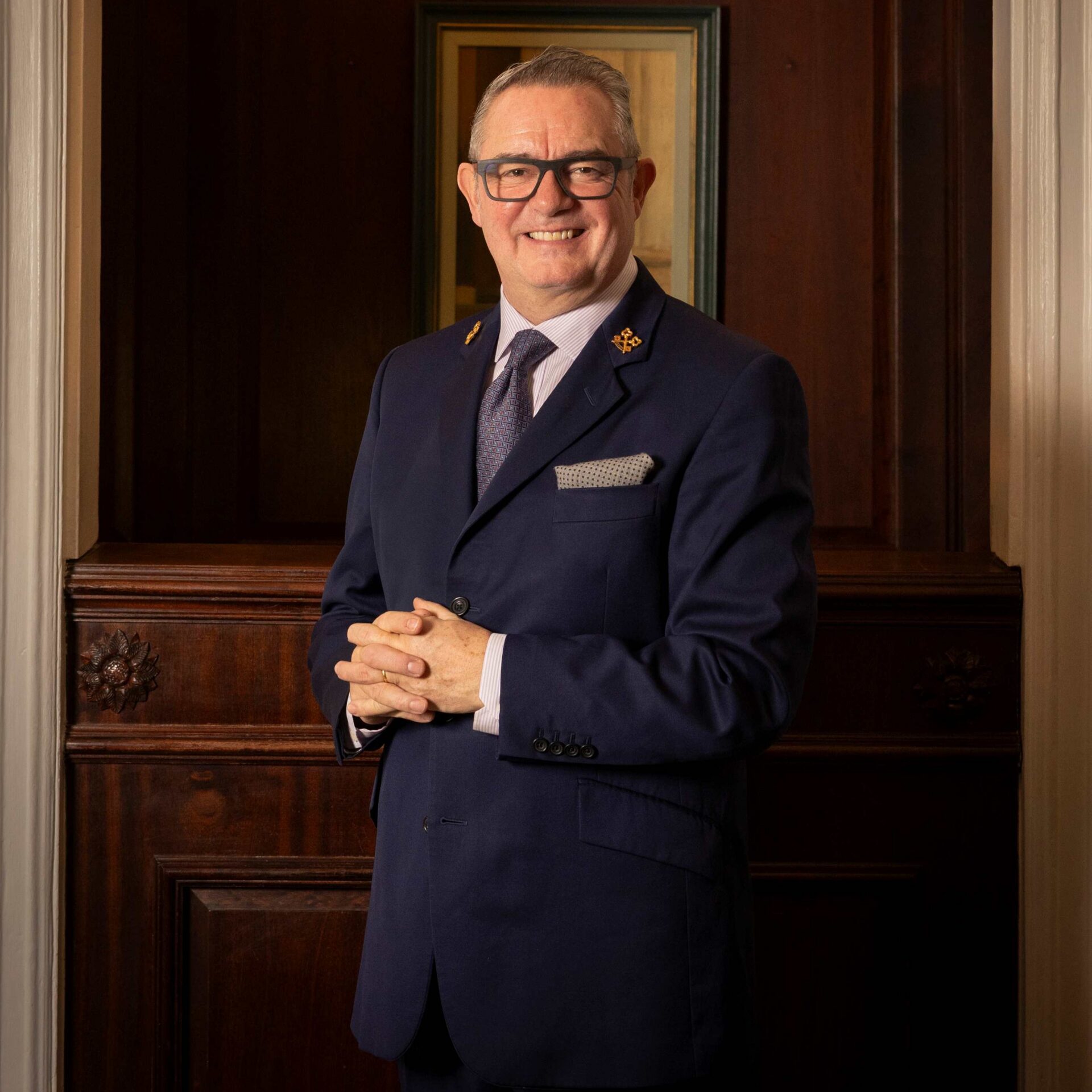 A man wearing glasses and a navy suit with a tie and pocket square stands smiling in front of a wooden wall, hands clasped together—exuding the professionalism often associated with premium chauffeur services.