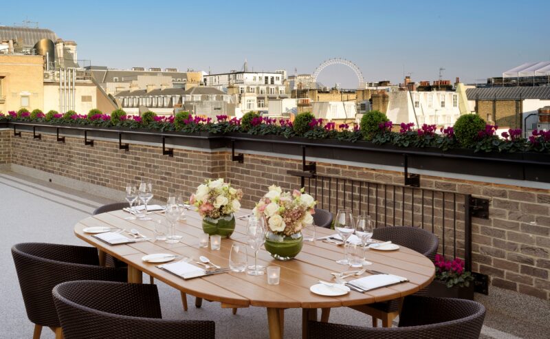 A rooftop dining area at The Stafford London features a wooden table set for six, floral centerpieces, and wicker chairs. The terrace overlooks city buildings, with the London Eye visible in the background under a clear blue sky.