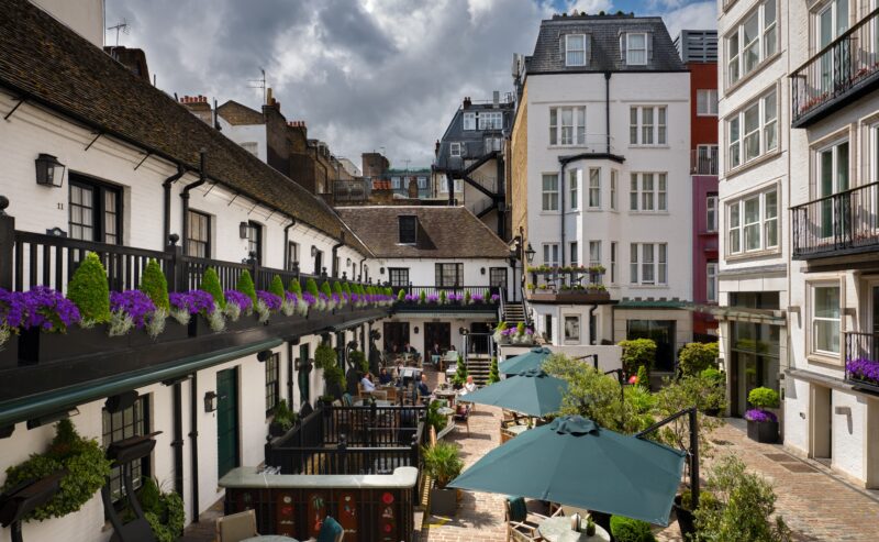 A charming courtyard at The Stafford London features outdoor tables, green umbrellas, potted plants, and purple flowers. White and colorful buildings with balconies surround guests dining and socializing under a partly cloudy sky.