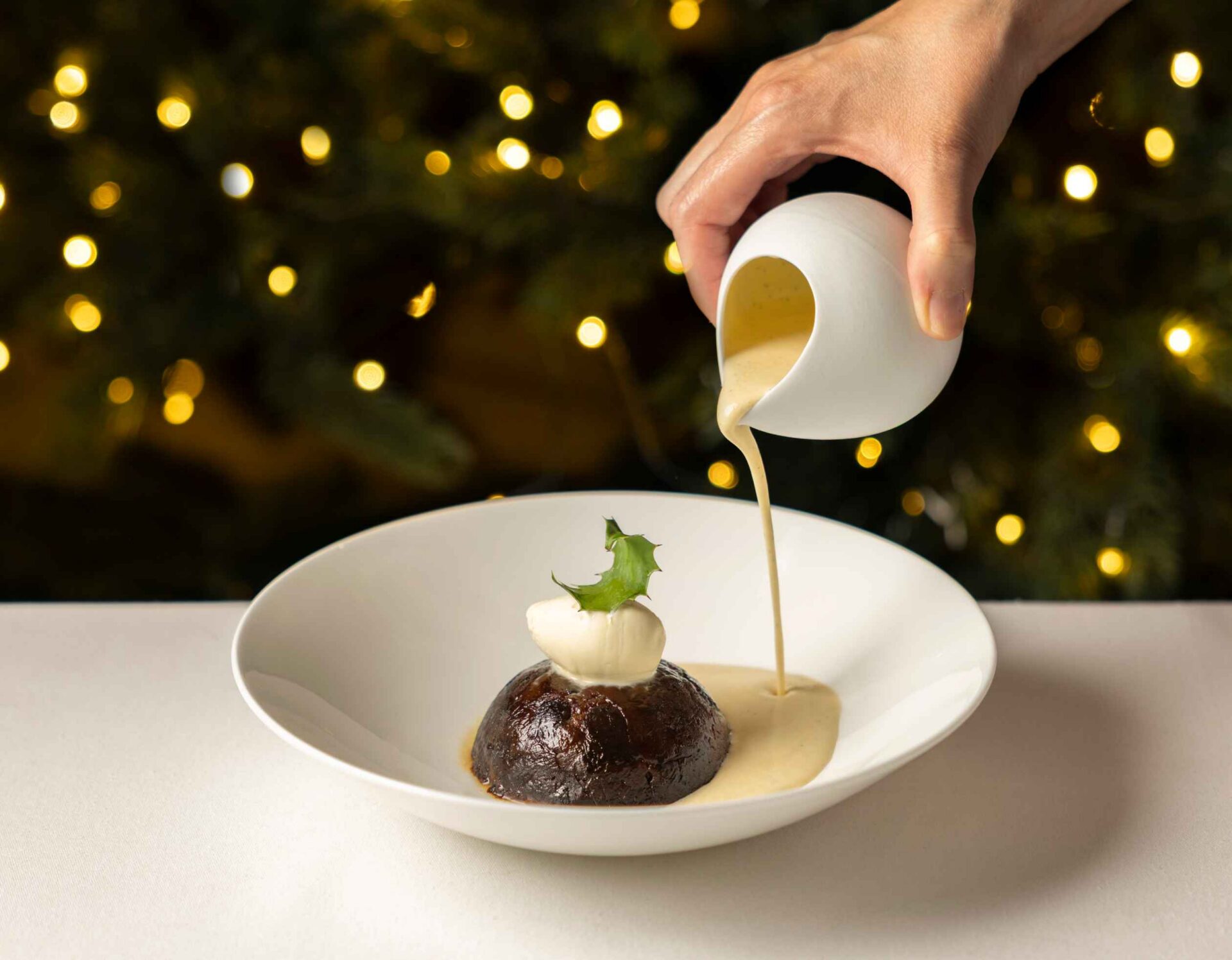 A hand pours creamy sauce from a small white jug onto a Christmas pudding topped with a dollop of cream and a holly leaf, with festive lights blurred in the background.