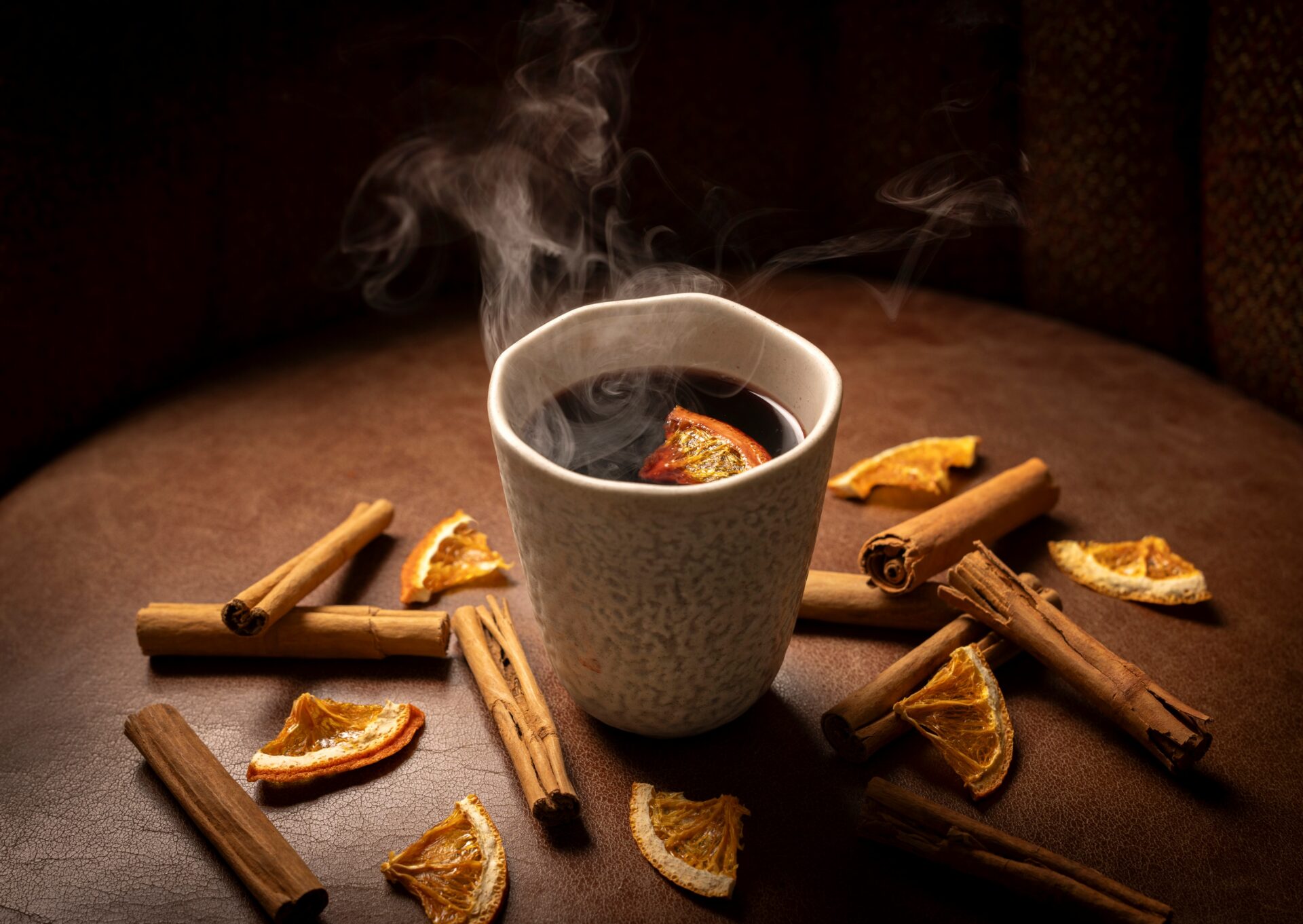 A steaming cup of mulled wine sits on a brown surface, surrounded by dried orange slices and cinnamon sticks, creating a warm and cozy atmosphere.