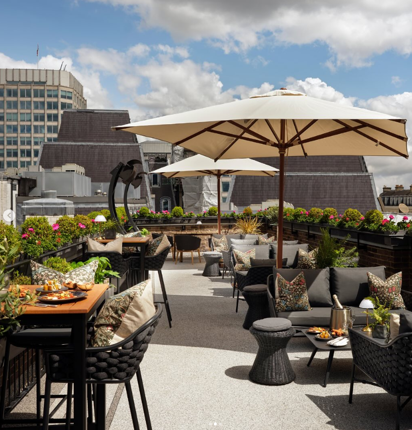 A rooftop terrace with tables, cushioned chairs, and large umbrellas. Potted plants line the edges, and food is served on the tables. City buildings and a partly cloudy sky are visible in the background.