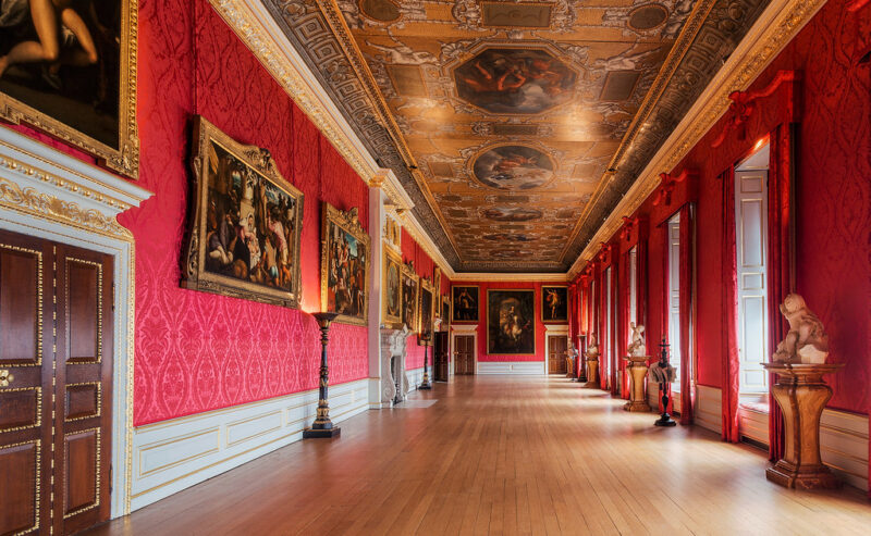 Ornate gallery with red walls, wooden floor, large paintings, sculptures, tall windows with red drapes, and a decorative ceiling featuring detailed artwork. The room is grand and well-lit by natural light.