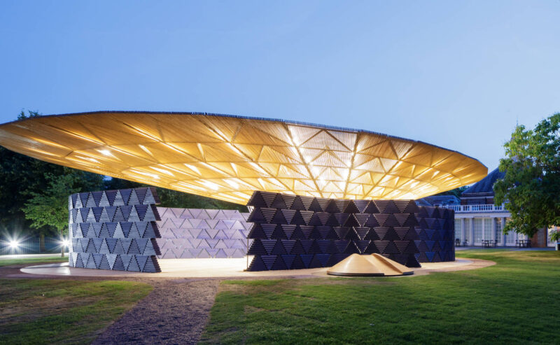 A modern pavilion with a large, illuminated, golden, geometric roof supported by dark, angled walls sits on a grassy lawn at dusk.