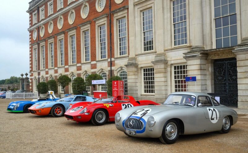 Five vintage race cars, each a different color, are parked in a row on gravel in front of a grand historic building with tall windows and ornate architecture.