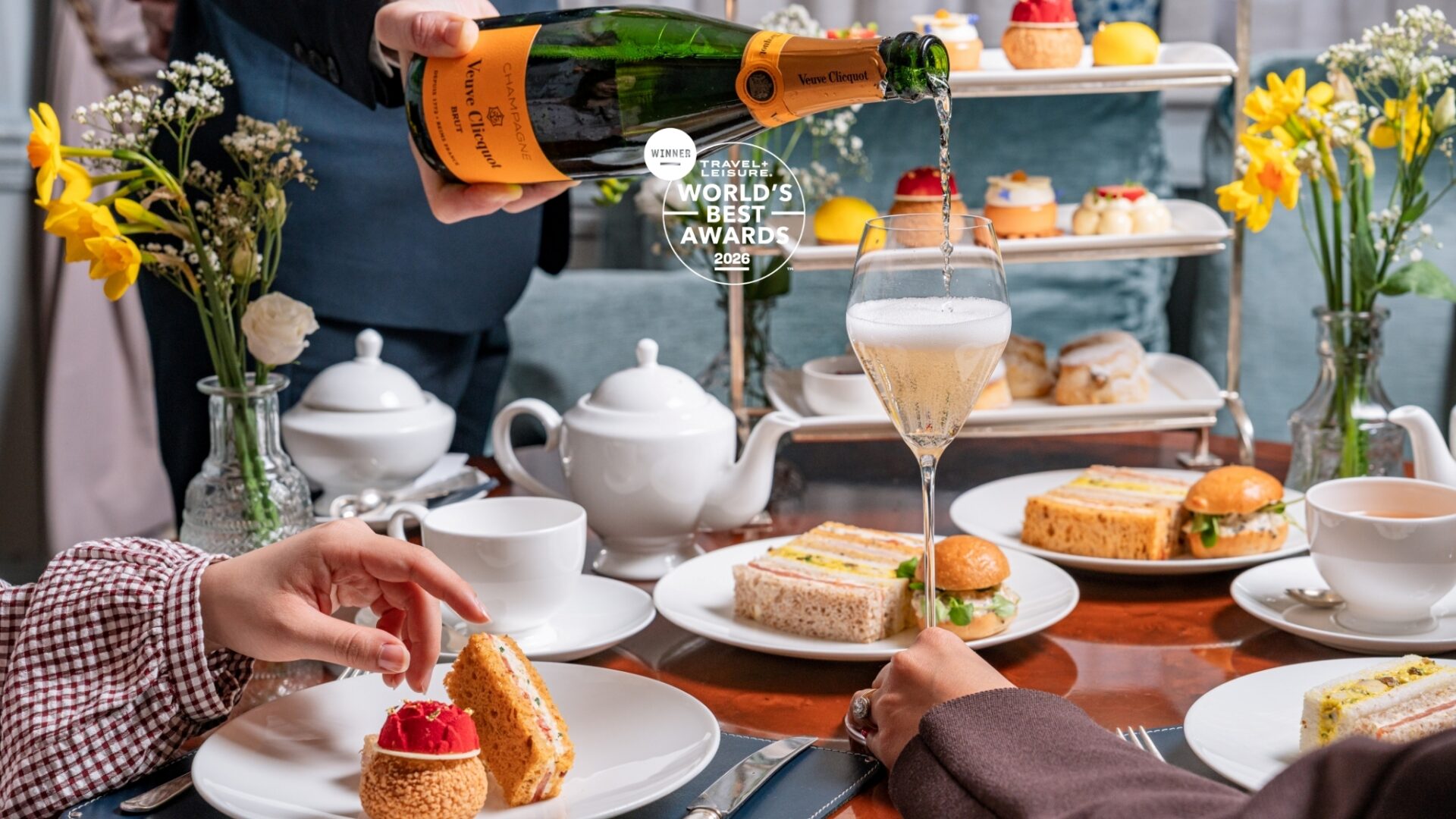 A person pours champagne into a glass at an elegant afternoon tea table at The Stafford London, surrounded by assorted pastries, sandwiches, and flowers. Two people are reaching for food while a three-tiered tray holds desserts in the background.