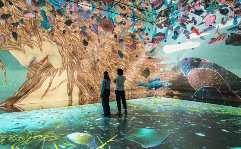 Two people stand in a room surrounded by colorful, immersive digital art projected on the walls, ceiling, and floor, creating a surreal, nature-inspired environment.