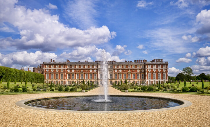 A round fountain in front of a large, ornate red-brick palace with many windows, surrounded by manicured gardens and a partly cloudy blue sky.