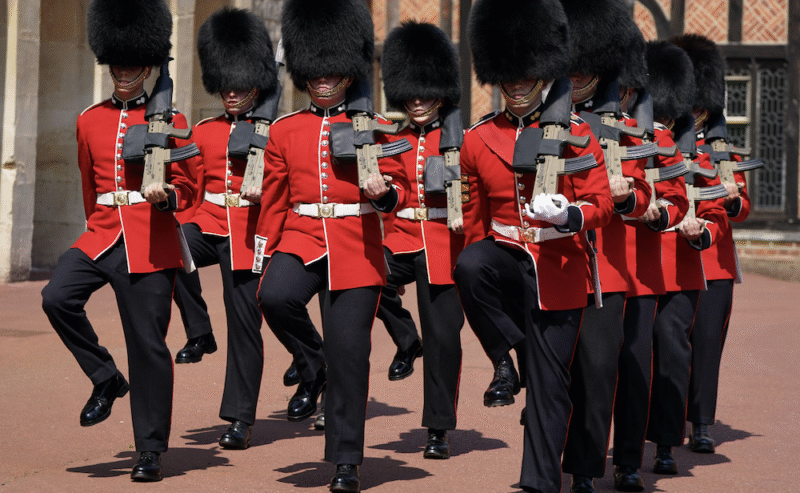 A group of British royal guards in red uniforms and tall black hats march in unison outside a historic building.