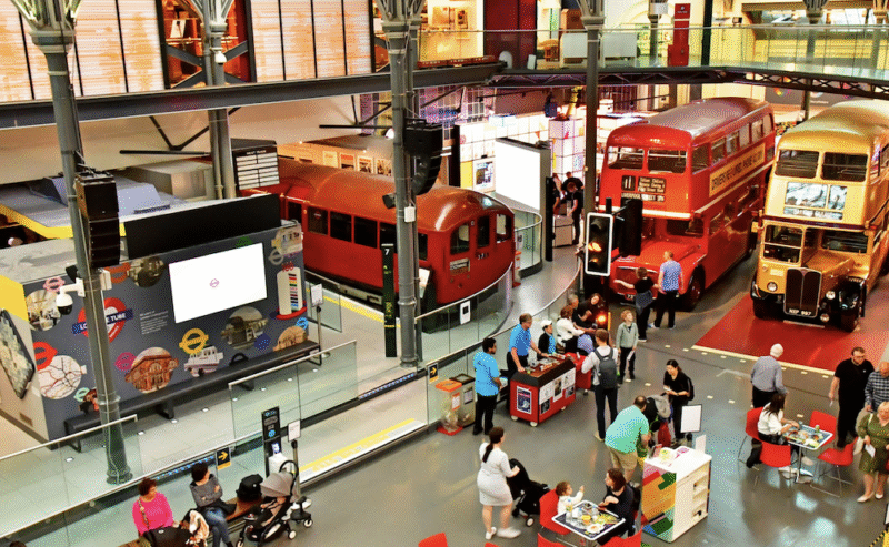 People explore a transport museum featuring a red subway car, two vintage double-decker buses, and informational exhibits in a bright, spacious hall. Some visitors sit at tables while others interact with displays.