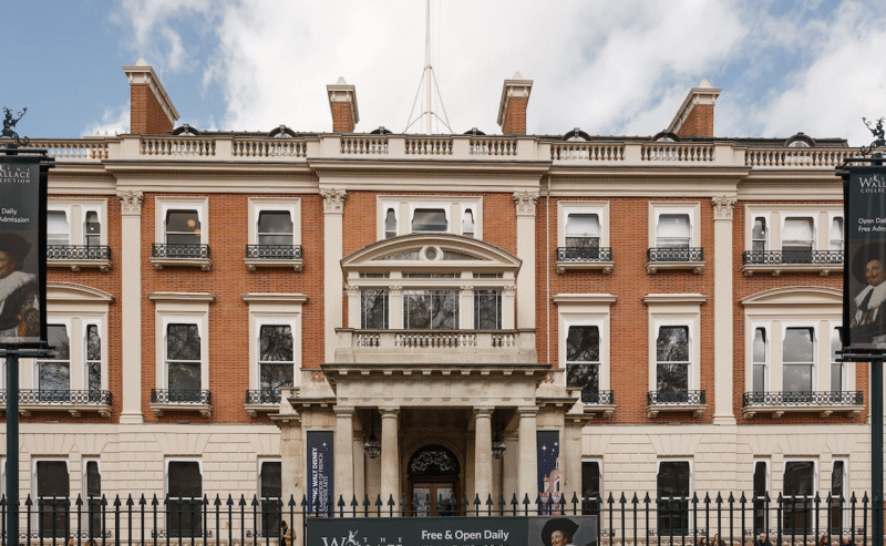 A large, historic brick museum building with columns at the entrance, signs reading The Wallace Collection, and banners displaying portraits, surrounded by a black iron fence.