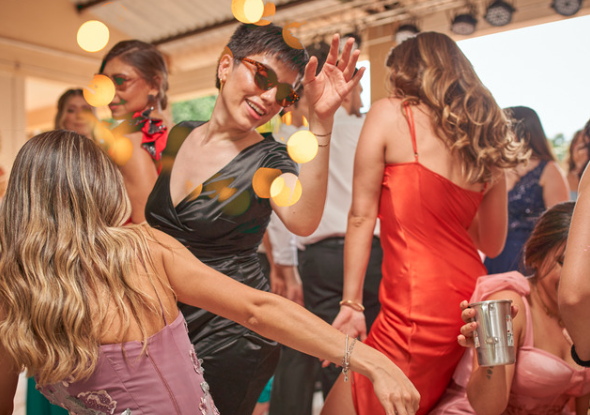 People in colorful dresses and sunglasses dance energetically at a lively indoor party, with yellow lights creating a festive atmosphere.