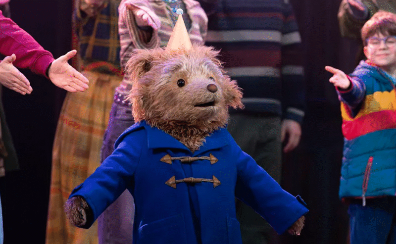 A bear wearing a blue coat and a party hat stands on stage, surrounded by people reaching their hands toward it.