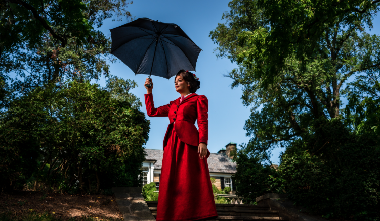 A person in a long red dress and bonnet stands outdoors holding a black umbrella, surrounded by green trees and shrubs with a house visible in the background.