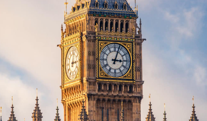 The clock tower of Big Ben in London, showing the time at 5:40. The ornate architecture is lit by sunlight, highlighting the gold and intricate details against a light sky.