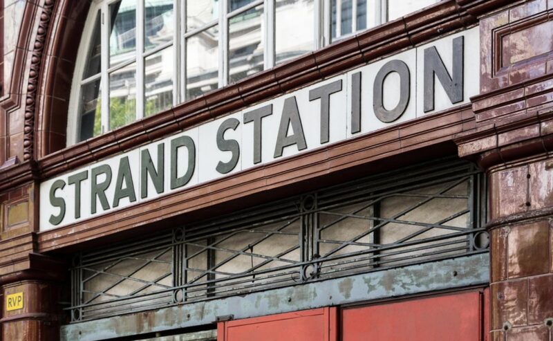 Close-up of the entrance to Strand Station, showing a large white sign with black letters above a gate and red-tiled exterior details.