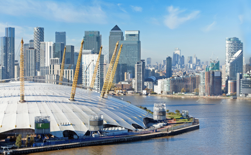 The O2 Arena in London by the River Thames, with yellow support beams. Modern skyscrapers and office buildings rise in the background, under a clear blue sky.
