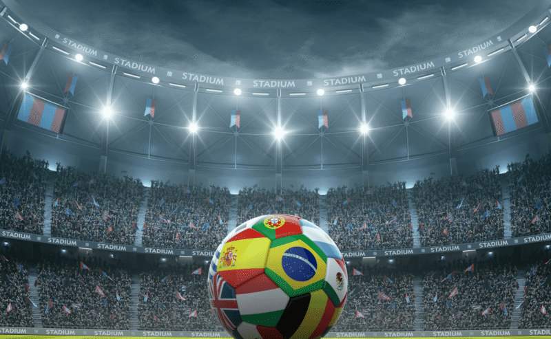 A soccer ball decorated with various national flags sits on a stadium field, surrounded by cheering crowds under bright stadium lights.