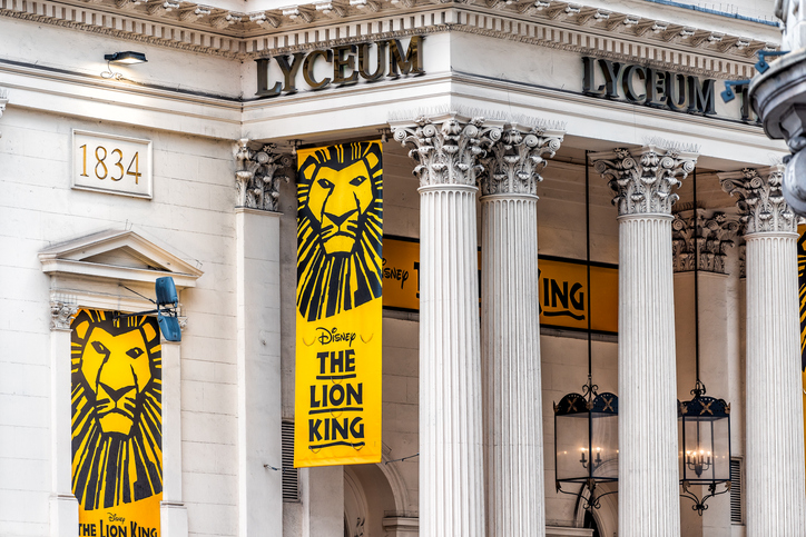 The exterior of the Lyceum Theatre, with yellow banners displaying Disney’s The Lion King logo and a lion’s face hanging between white columns.