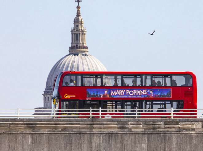 A red double-decker bus with a Mary Poppins ad crosses a bridge in front of St. Pauls Cathedral dome in London, with a bird flying in the clear sky.