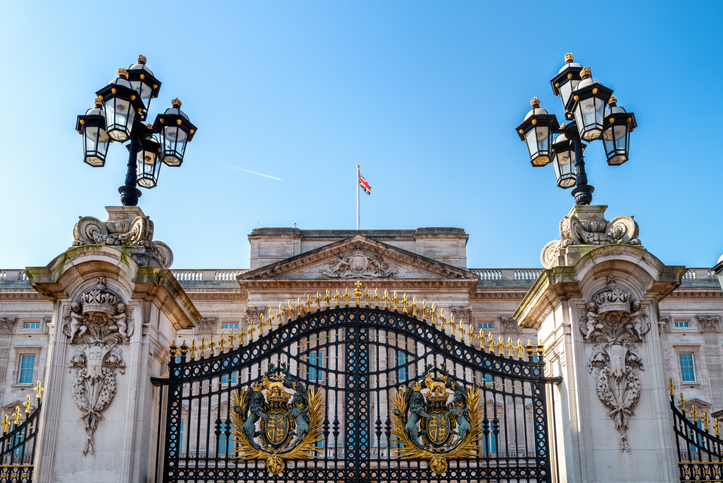 Buckingham Palaces ornate front gates with gold details, lanterns, and the British flag flying atop the building, set against a clear blue sky.