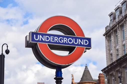 A London Underground sign with a red circle and blue bar reading Underground against a partly cloudy sky and historic buildings.