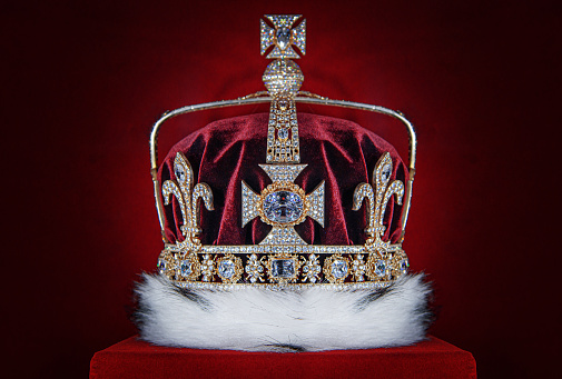 A jeweled crown with gold, diamonds, and a red velvet cap, decorated with white fur trim, sits against a deep red background.
