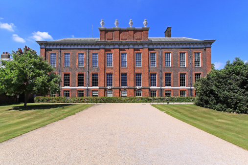 A symmetrical red-brick building with tall windows, a decorative roofline, and statues on top, surrounded by green lawns, trees, and a gravel path under a clear blue sky.
