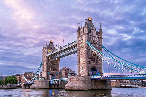 The Tower Bridge in London, England, is shown at sunset with its twin towers and blue suspension elements reflected in the River Thames under a cloudy sky.