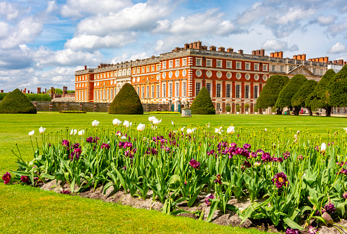 Red-brick palace with many chimneys, manicured lawns, trimmed hedges, and colorful tulips blooming in the foreground under a partly cloudy sky.