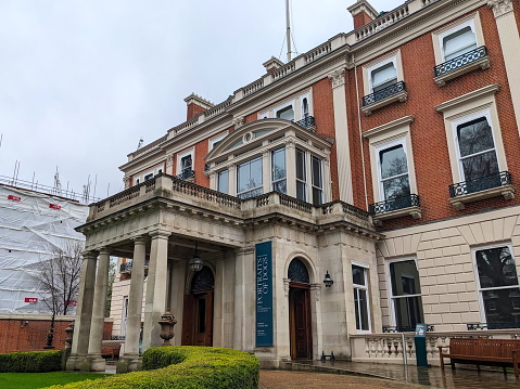 A red-brick and white stone building with columns at the entrance, featuring large windows and a banner. The surrounding area is wet and a nearby structure is covered in scaffolding.
