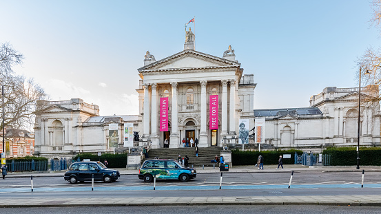 Neoclassical building with columns and banners reading Tate Britain and Free for All. People walk up the front steps; cars and a taxi are on the street in front. Trees frame the scene under a clear sky.