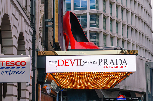 Theater marquee with a large red high heel above it reads, “The Devil Wears Prada: A New Musical.” A Tesco Express sign is visible to the left.