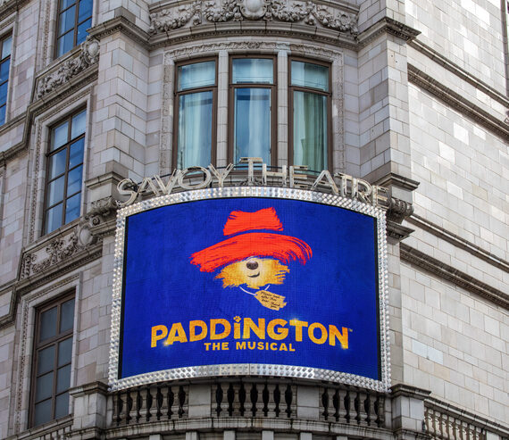 A theater sign reads Paddington The Musical with an image of a bear in a red hat and coat, displayed on the Savoy Theatres exterior.
