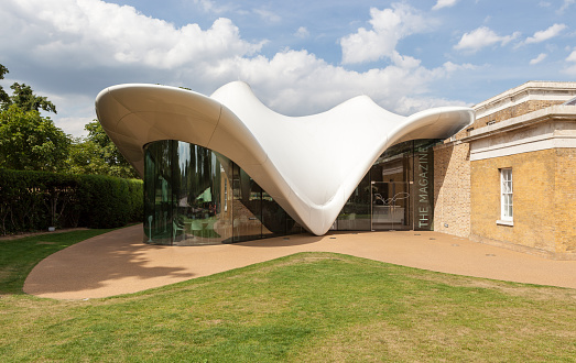 A modern building with a white, wave-shaped roof and glass walls, attached to a traditional brick structure. The entrance reads THE MAGAZINE. The building is surrounded by grass and hedges under a partly cloudy sky.