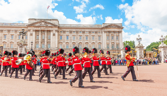 A marching band of guards in red uniforms and bearskin hats performs in front of Buckingham Palace, with a crowd watching behind gates on a sunny day.