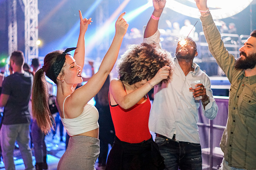 A group of people dance and celebrate at a lively outdoor party at night, some holding sparklers and drinks, with colorful lights in the background.