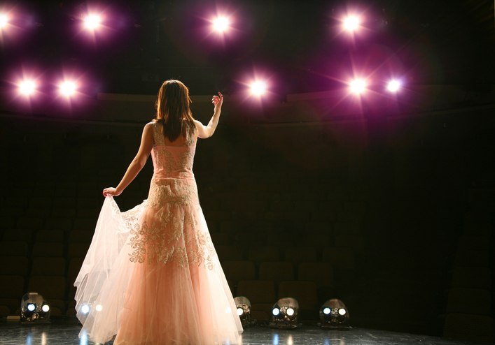 A woman in a long, elegant pink dress stands on stage facing bright lights, holding part of her skirt as she gestures with one arm, performing to an empty, dimly lit theater.