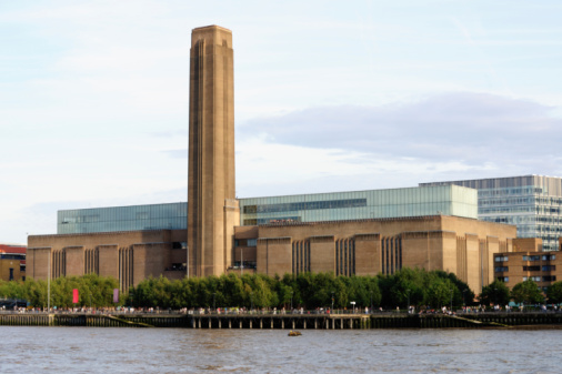 A large brick building with a tall central chimney, the Tate Modern art museum, stands beside the River Thames with trees lining the waterfront under a partly cloudy sky.