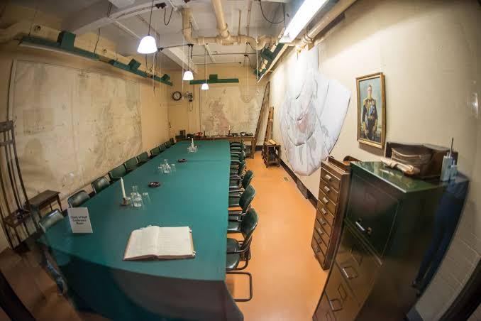 A long conference table with green chairs in a vintage, bunker-like room. Maps, a portrait, and filing cabinets are on the walls. A large open book and glass bottles sit on the table. The lighting is bright.