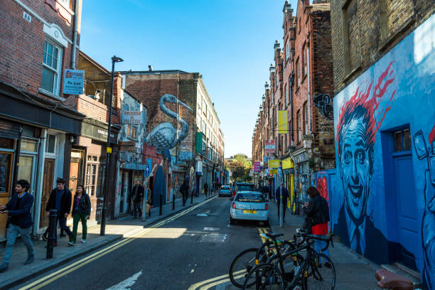 A lively city street lined with brick buildings, colorful murals, parked cars, bicycles, and people walking on the sidewalks under a clear blue sky.