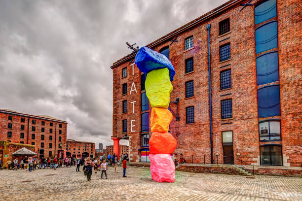 A colorful vertical sculpture of stacked rocks stands in front of a red brick building with blue windows on a cloudy day. People gather around the artwork in a cobblestone courtyard.