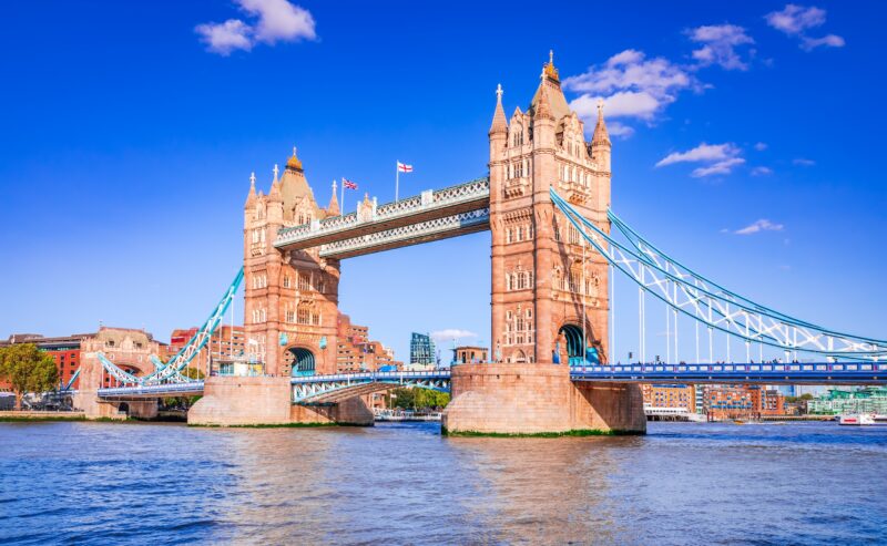 Tower Bridge in London on a sunny day, with blue sky and scattered clouds, spans the River Thames. The iconic twin towers and suspension cables are clearly visible, with city buildings in the background.