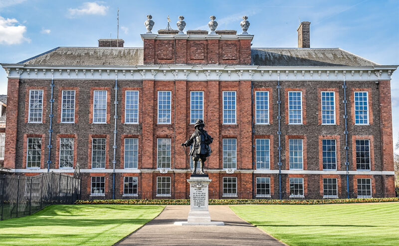 A large red-brick building with many windows and a statue in front, centered on a neatly mowed lawn under a blue sky.