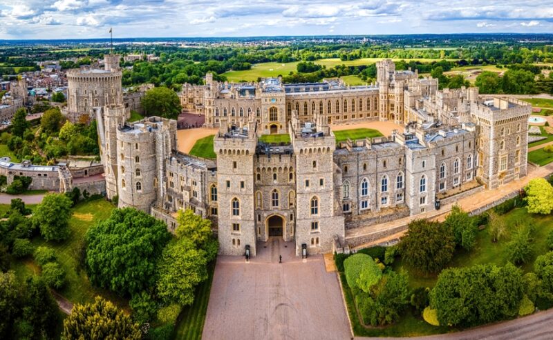 A panoramic aerial view of Windsor Castle, showcasing its stone walls, towers, and green surroundings under a partly cloudy sky.