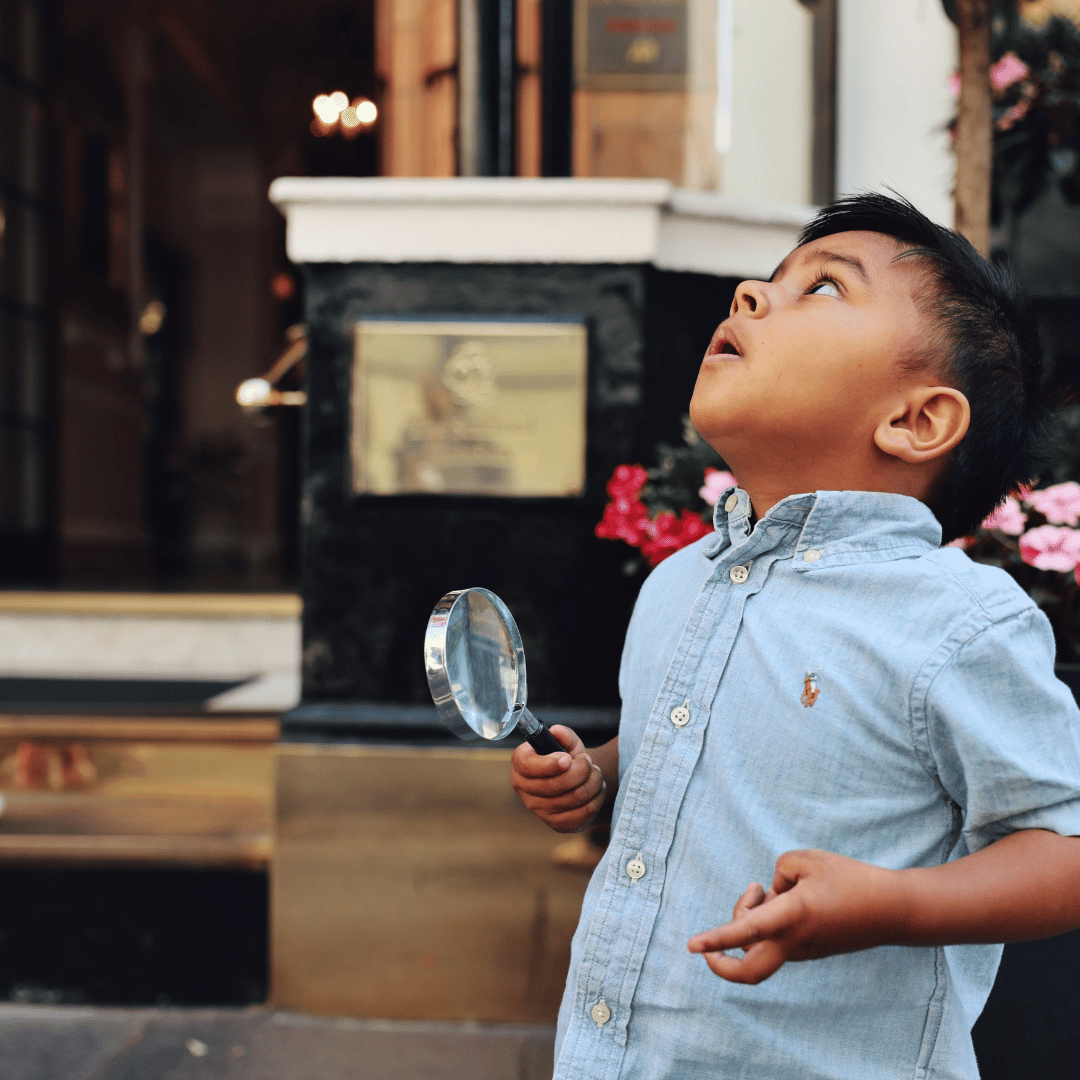 A young boy holding a magnifying glass looks up with curiosity outside a building, wearing a light blue button-up shirt. Flowers and a plaque are visible in the background, hinting at welcoming room offers inside.