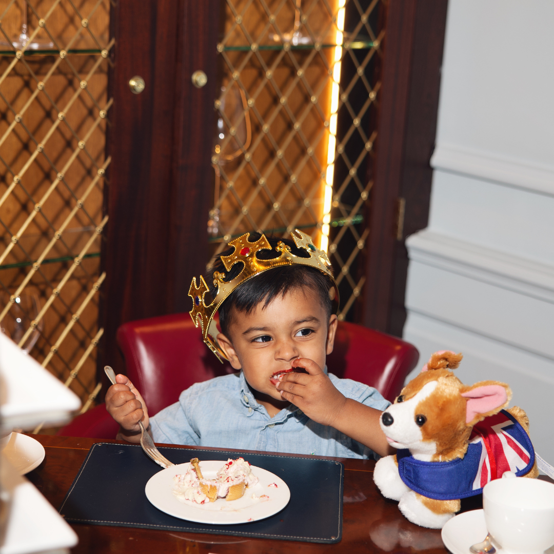 A young child wearing a crown enjoys afternoon tea, eating cake at a table while sitting in a red chair next to a stuffed dog toy dressed in a Union Jack outfit. Shelved cabinets are in the background.