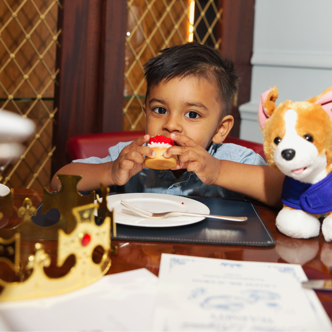 A young boy sits at a table enjoying a cupcake during afternoon tea. A toy dog and a gold crown rest on the table beside him as he looks towards the camera in a cozy indoor setting.